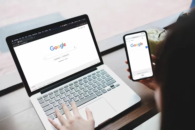 A person sits at a laptop with Google search displayed on the browser, while simultaneously searching on a phone. This image signifies AI ecommerce integration, showcasing how artificial intelligence algorithms analyze search queries across devices to provide tailored product suggestions and enhance the shopping experience.
