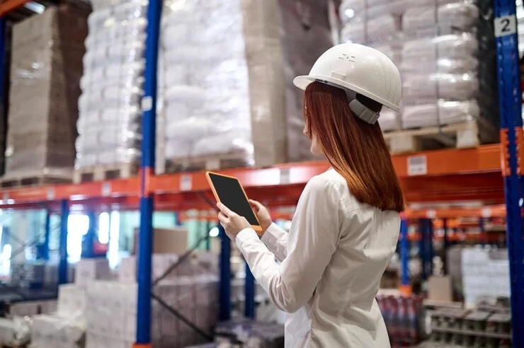 A woman glances at her device while walking into a warehouse, representing AI ecommerce integration. This image symbolizes the use of artificial intelligence to streamline inventory management processes. Additionally, it hints at shipping strategies, suggesting the role of AI in optimizing logistics and fulfillment for efficient delivery.