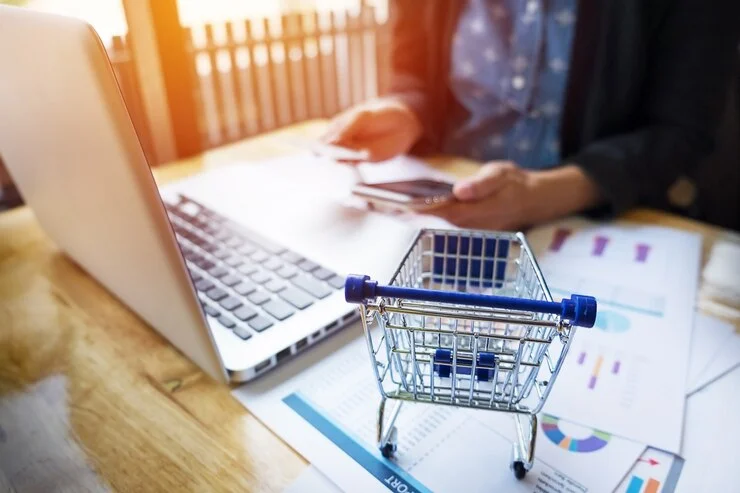 A person sits at a laptop with a phone in hand, accompanied by a miniature shopping cart. This image illustrates AI ecommerce integration, showcasing how artificial intelligence enhances online shopping experiences by analyzing data and suggesting personalized product recommendations to users.