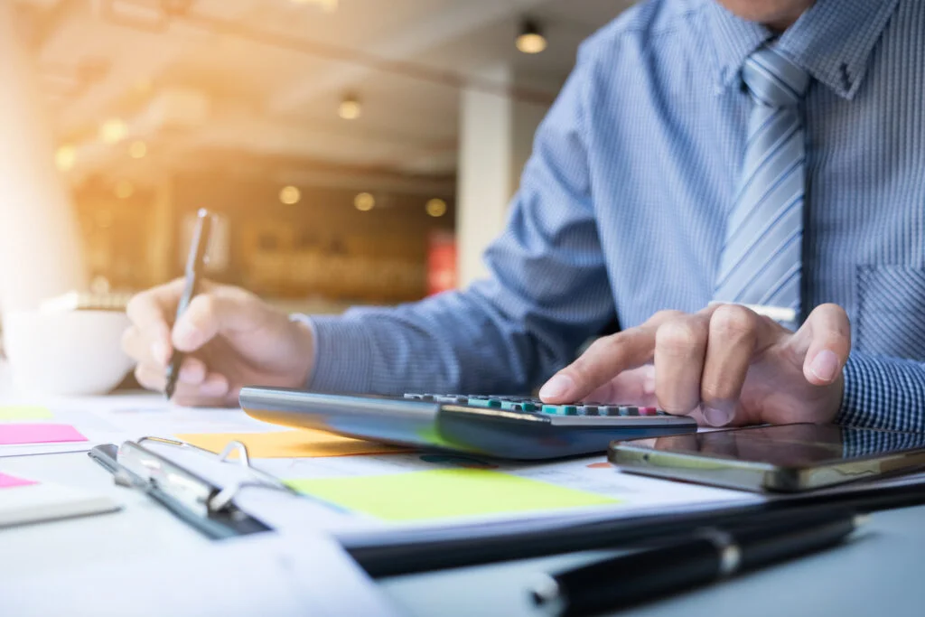 A person multitasking with a calculator and writing tools, symbolizing the active navigation of dynamic pricing strategies amidst essential eCommerce updates.